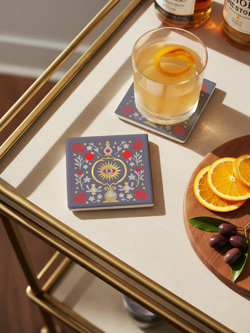 A square ceramic drink coaster with a smooth, matte charcoal grey finish. The surface features a symmetrical, intricate design in metallic gold, deep garnet red, and light grey. In the center is a golden All-Seeing Eye with a red iris, radiating light. The eye is framed by a golden circle and flanked by botanical elements, including red pomegranates and grey flowers with gold details. The design has a folk art, gothic botanical style.